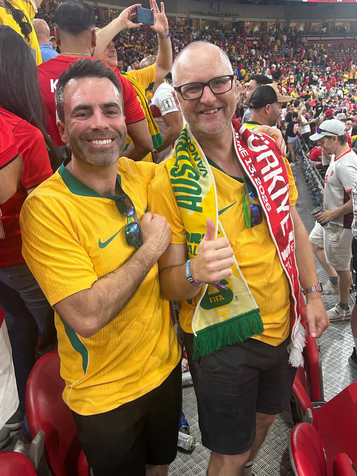 A dark-haired man and a man in glasses smile while sitting among a huge crowd in a football stadium.