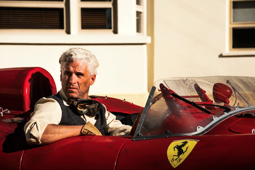 A film still of Patrick Dempsey, with white hair, seated in a red Ferrari, with a determined expression.