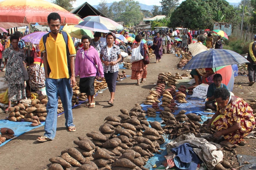 People walk past sweet potatoes laid out on the ground at a colourful market