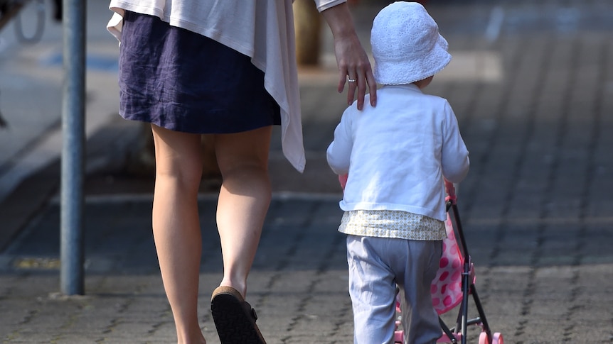 a woman holds the back of a toddler who is pushing a stroller along a footpath