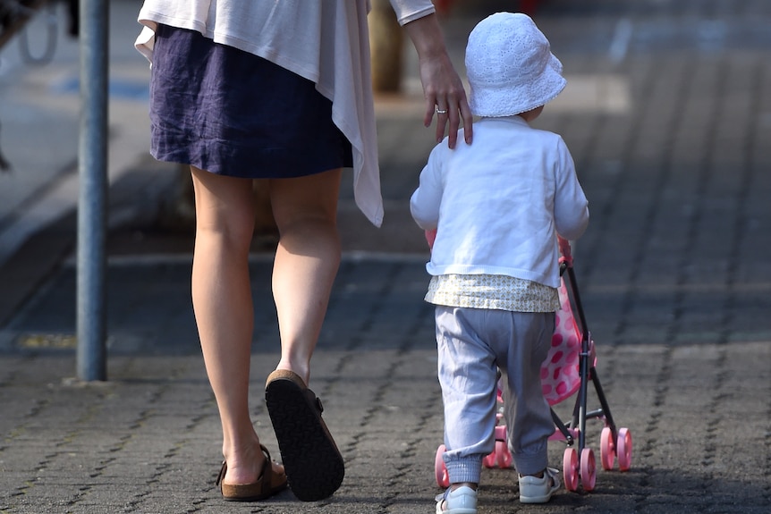 a woman holds the back of a toddler who is pushing a stroller along a footpath