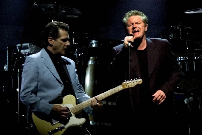 One man in a blue jacket on stage plays a cream-coloured electric guitar while another man in darker jacket sings
