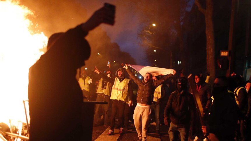 A figure is silhouetted against a bonfire, while in the background people wearing yellow vests (one carrying a French flag) yell
