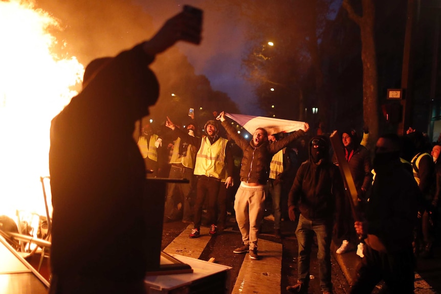 A figure is silhouetted against a bonfire, while in the background people wearing yellow vests (one carrying a French flag) yell