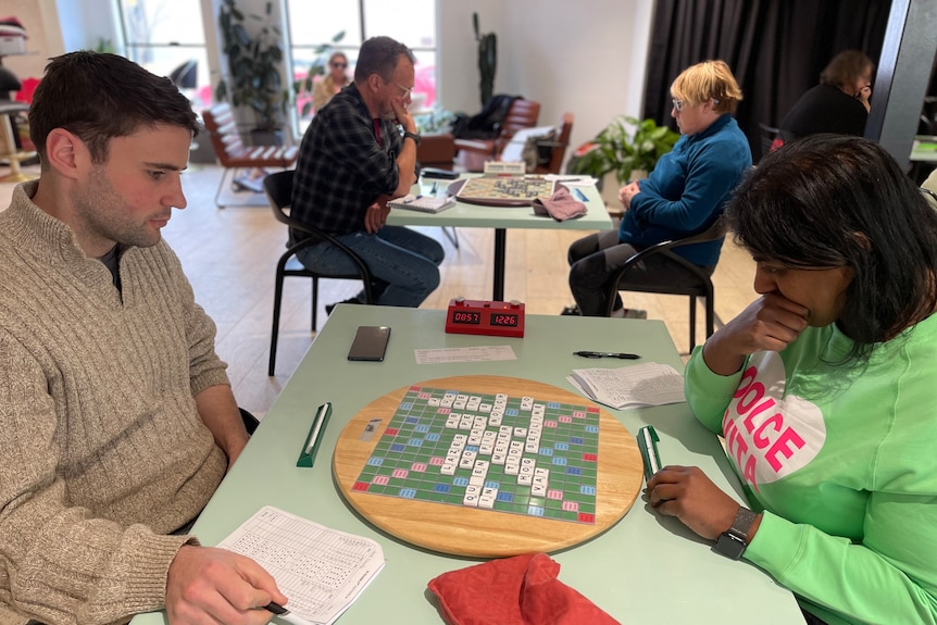 Two people sit opposite each other at a table with a Scrabble board between them 