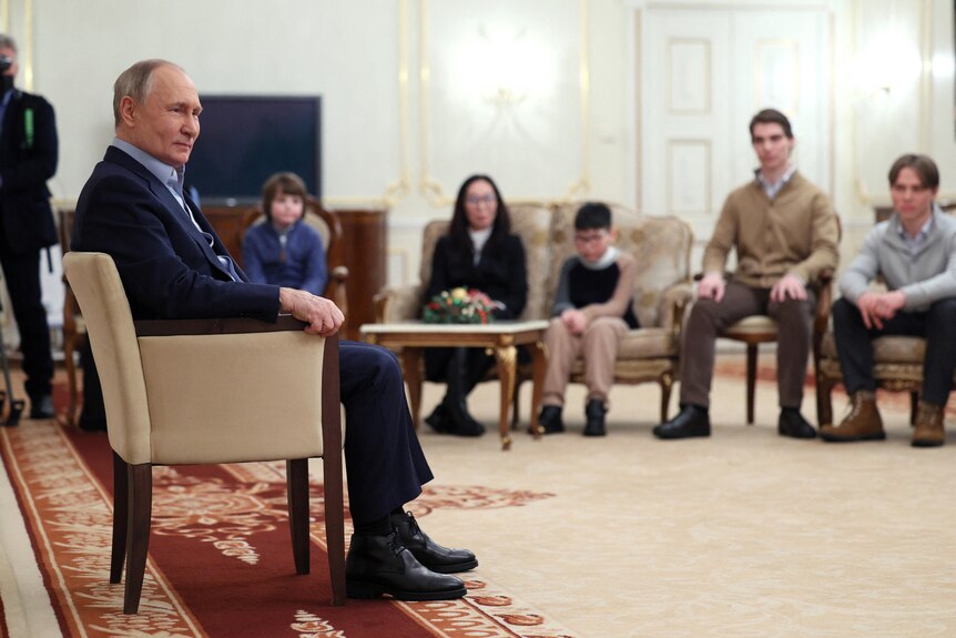 Vladimir Putin sits in a chair in a room where relatives of killed Russian servicemen sit nearby