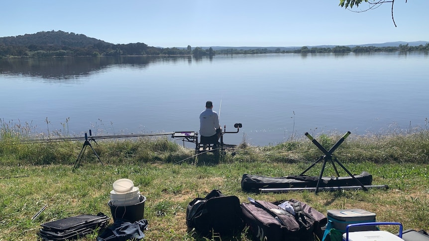 A man sits on the shore of lake burley griffin fishing. There is fishing paraphernalia in the foreground.