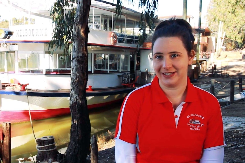 A young woman in a red and white top stands on the river bank beside a paddle steamer.