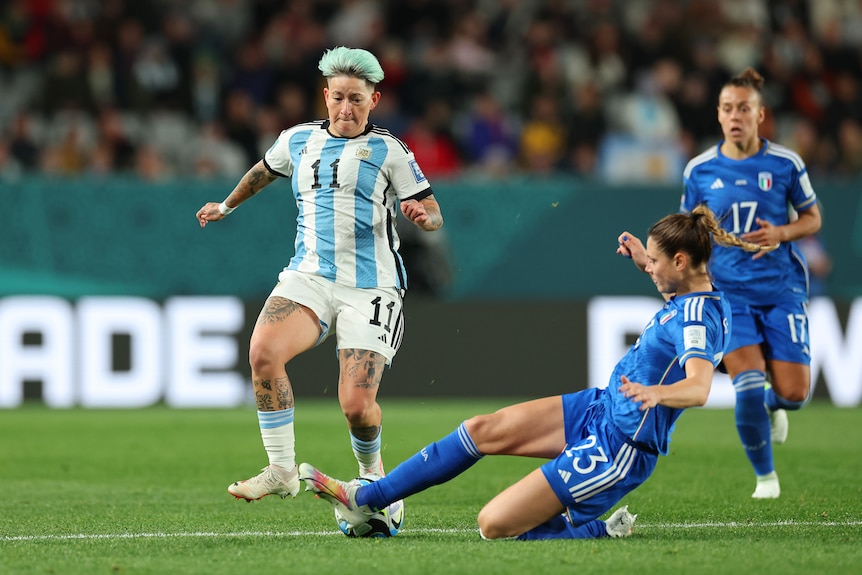 A women's footballer for Argentina looks down as an Italian defender slides in to tackle her and get the ball.