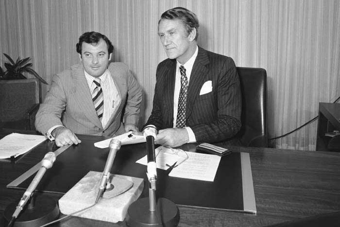 A black-and-white photo of two men in suits sitting at a desk.