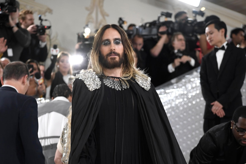 Jared Leto wears a long black cape with a black outfit underneath. The shoulders of the cape have silver jewels.