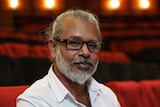 47-year-old Sri Lankan man with grey beard, moustache and long hair pulled back, wearing a white shirt, sitting in theatre.