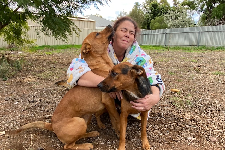 A woman with her arms wrapped around two large dogs squatting outside.