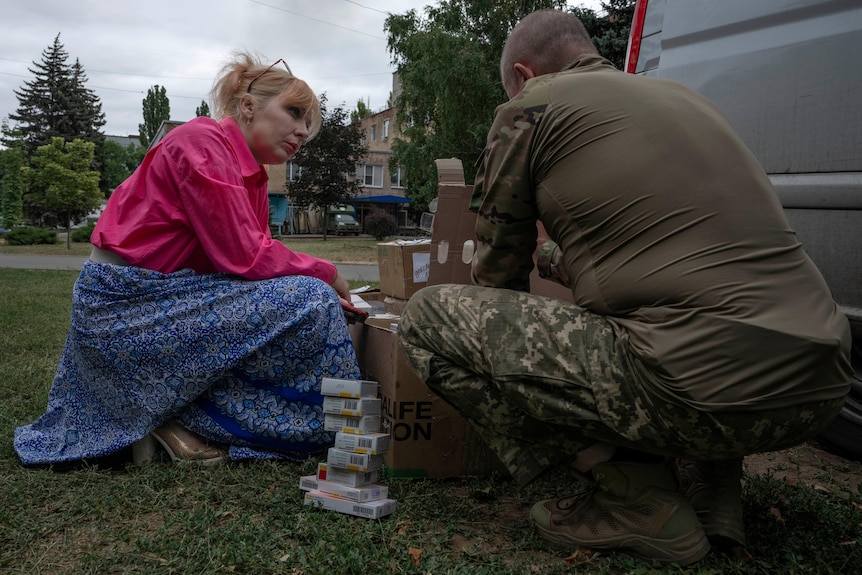 Medic volunteer Nataliia Voronkova dressed in bright pink and blue, kneels next to a soldier in camouflage uniform. 