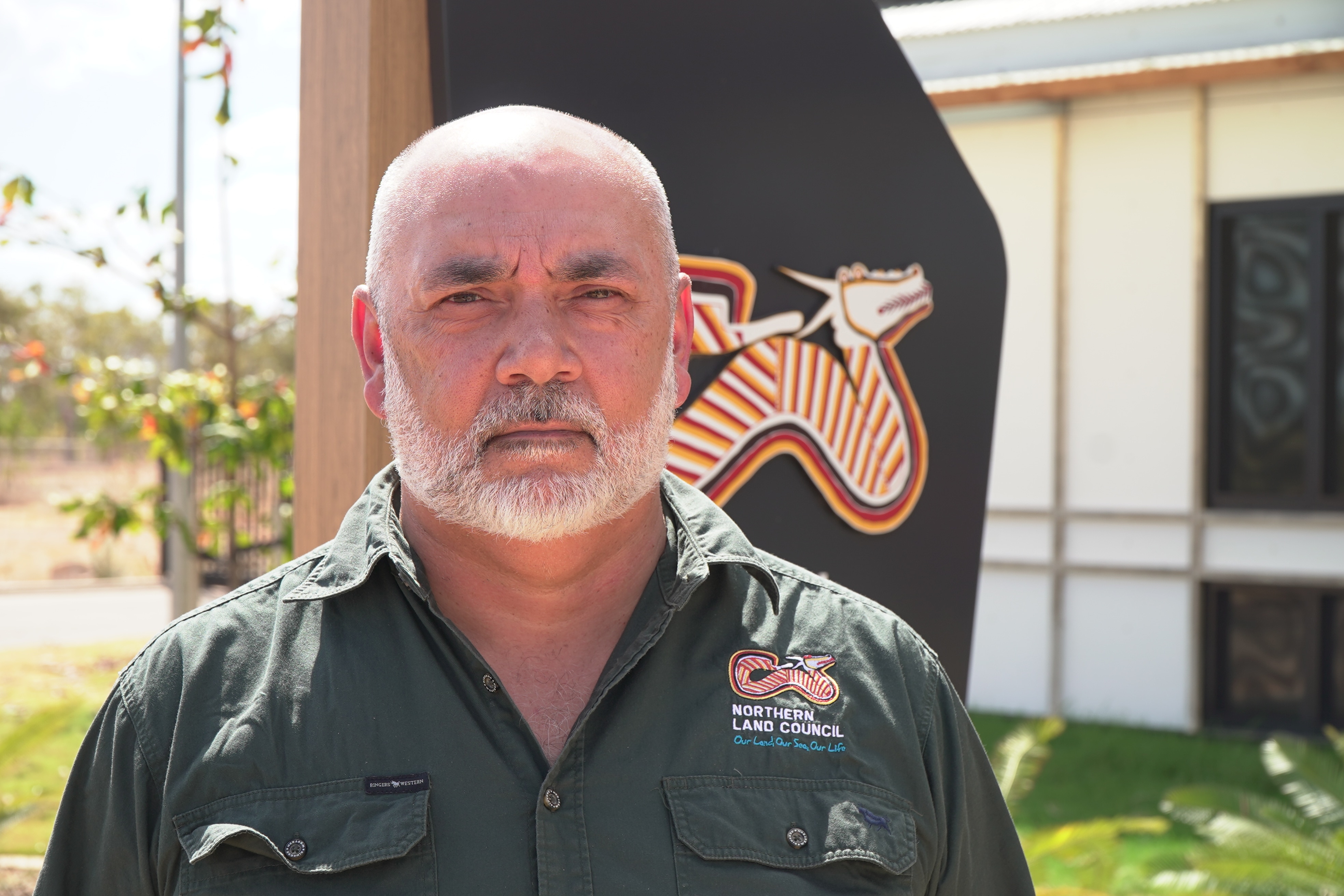 A close up shot of a pale man with a gray beard and moustache, wearing a khaki button up workers shirt.