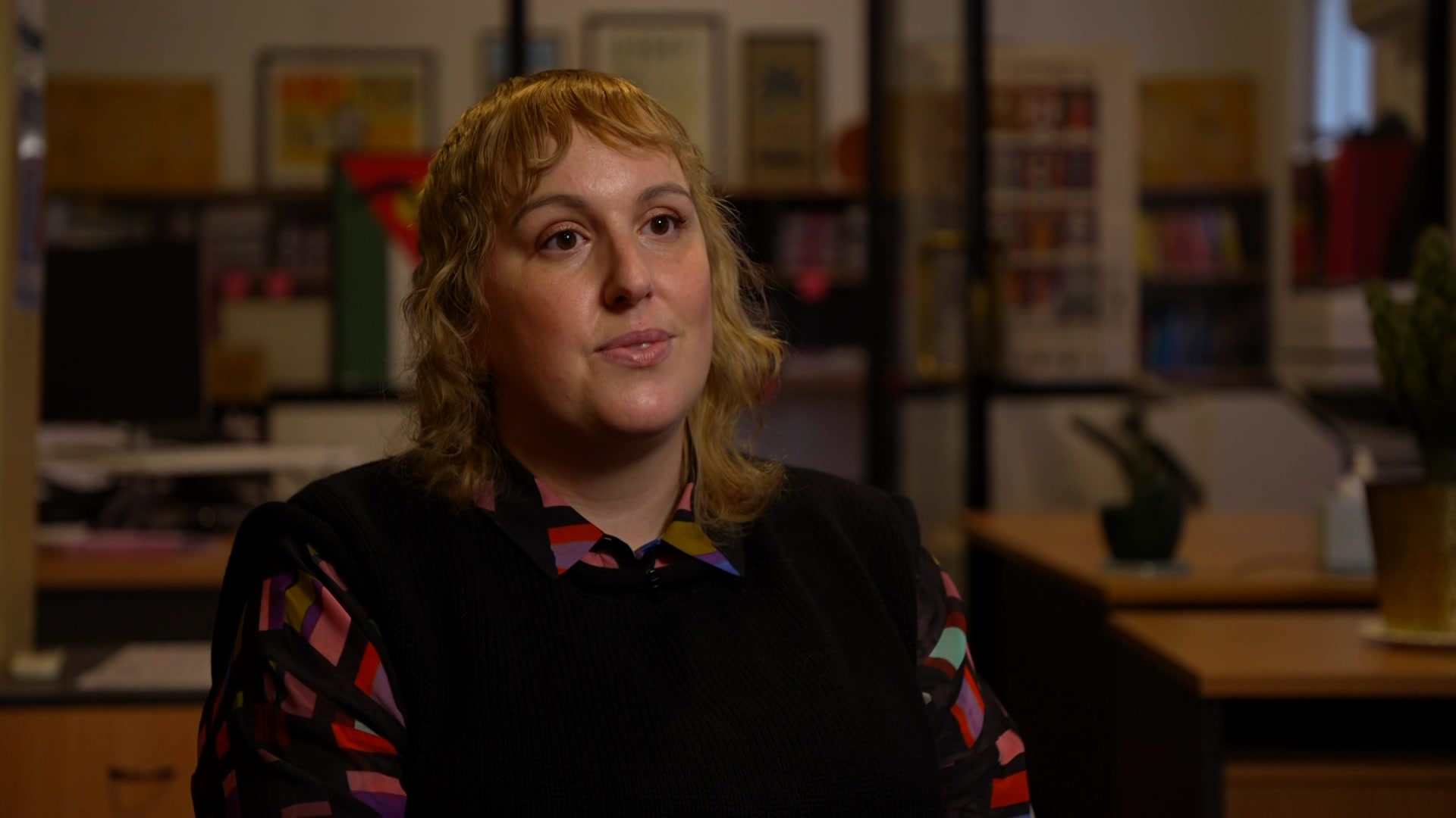 A blond curly hair woman wearing a shirt and a vest sitting in an office.