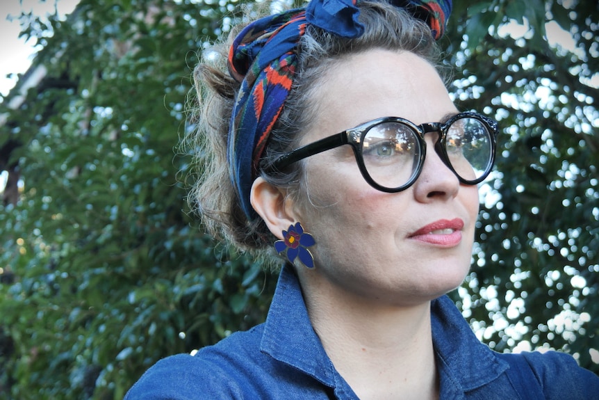 A headshot of a woman with a blue shirt and headscarf. She is also wearing glasses and is outdoors.