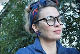 A headshot of a woman with a blue shirt and headscarf. She is also wearing glasses and is outdoors.