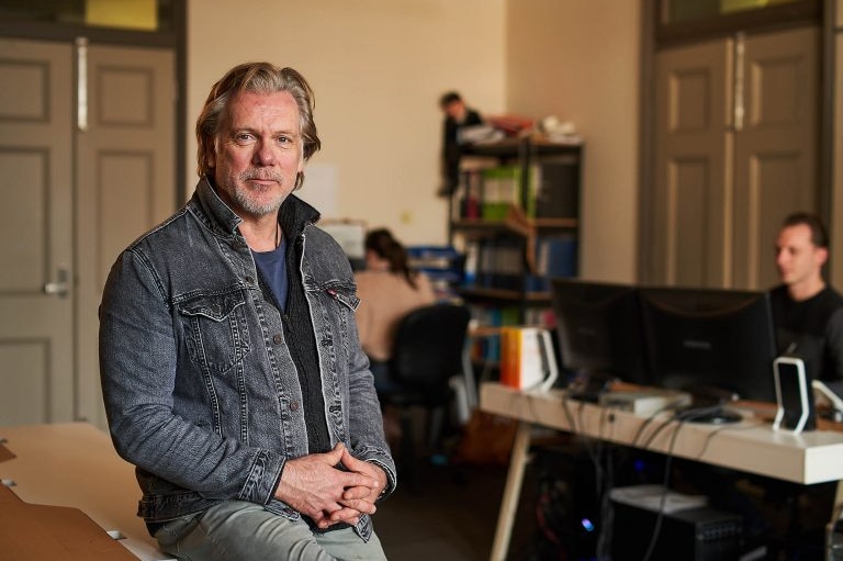 A man sitting on a table with hands linked looing at the camera.