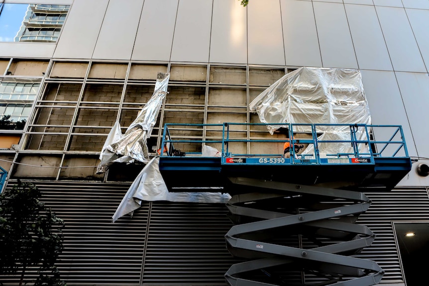 Building exterior where several panels are being removed by worker on scissor lift, with tarp and covering blowing in wind.