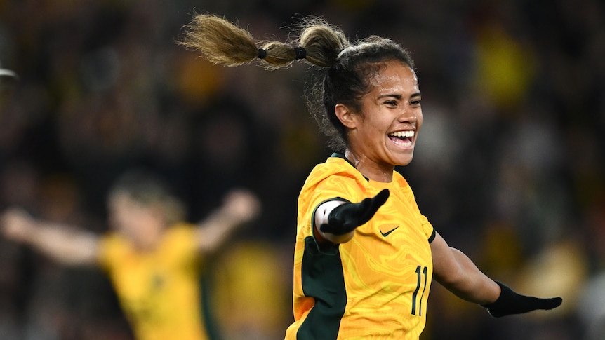 A female footballer in a gold Australia jersey smiles and celebrates with her arm extended, celebrating a goal