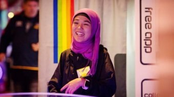 Rahila smiles as she looks at someone opposite her. She wears a purple head scarf and stands in front of a rainbow curtain.