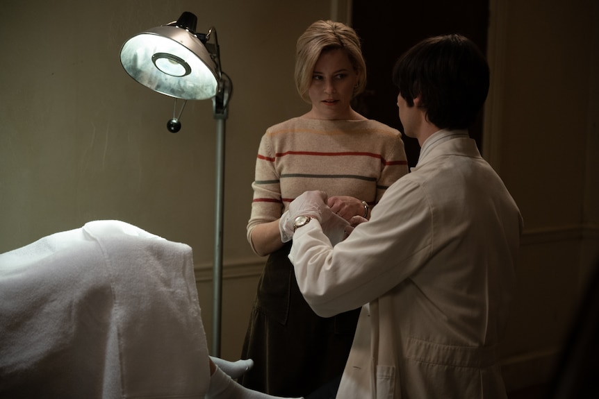 A man in a lab coat hands a medical implement to a middle-aged blonde woman, in a makeshift operating theatre