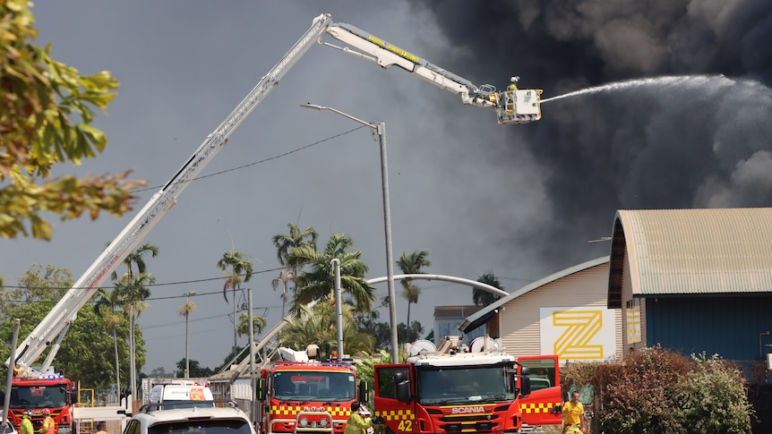 A crane shoots water into a plume of black smoke. Red fire trucks are seen down below. 