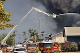 A crane shoots water into a plume of black smoke. Red fire trucks are seen down below. 