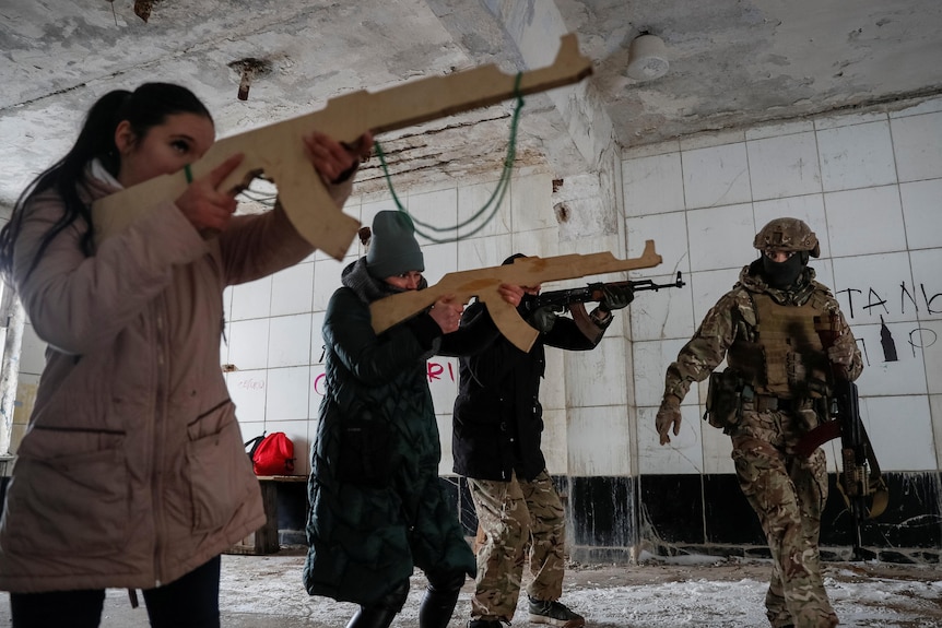 Several people stand in a disused building holding timber replicas of firearms. A person in full military gear walks past