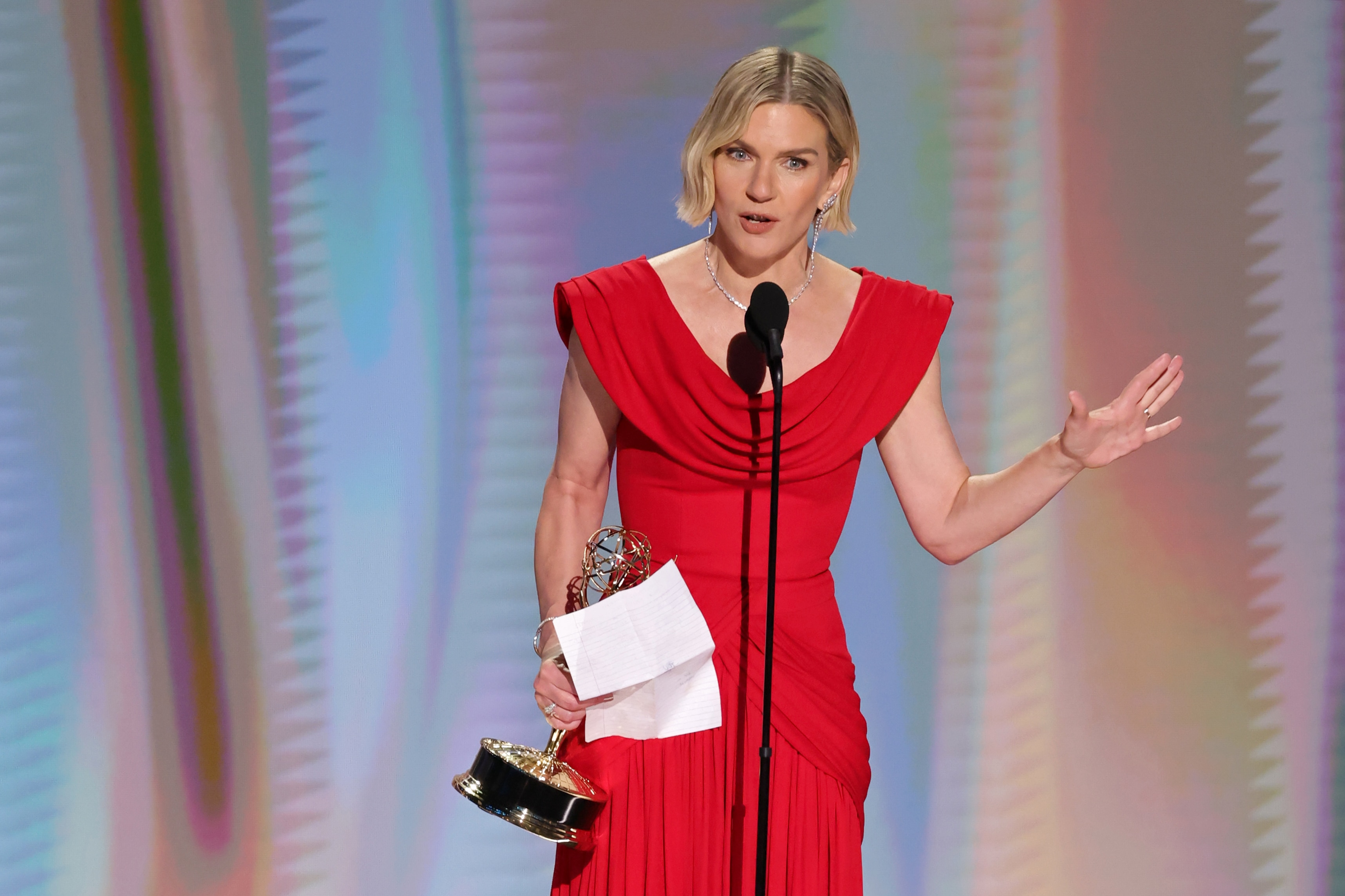 Rhea Seehorn wears a red gown and holds an award and gestures with her arm as she talks on stage behind a microphone.
