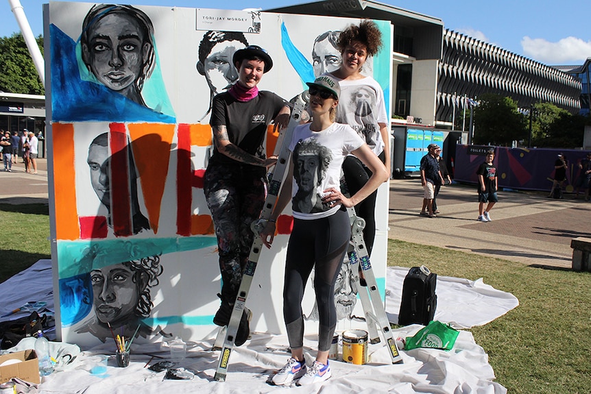 Three female street artists posing on a ladder beside a mural at South Bank, Brisbane.