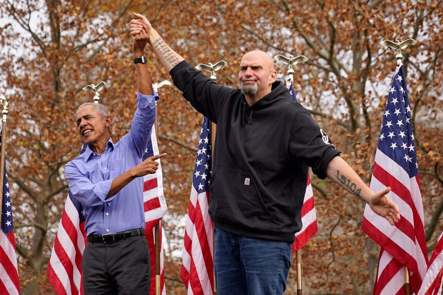 Barack Obama is seen holding John Fetterman's hand in the air, pointing towards him.