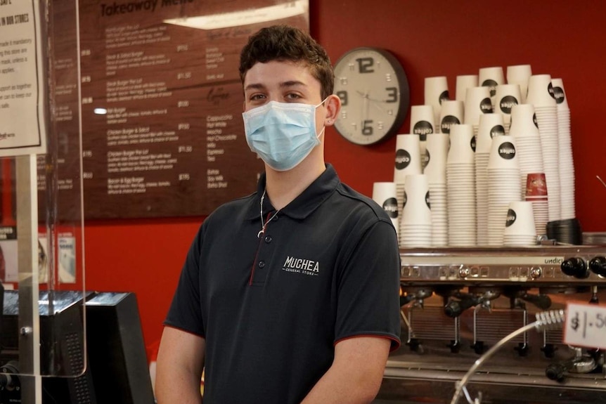 Muchea supermarket worker Mitchell White pictured wearing a mask with coffee cups stacked behind him.