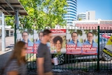 Two people walking past fence with election posters. 