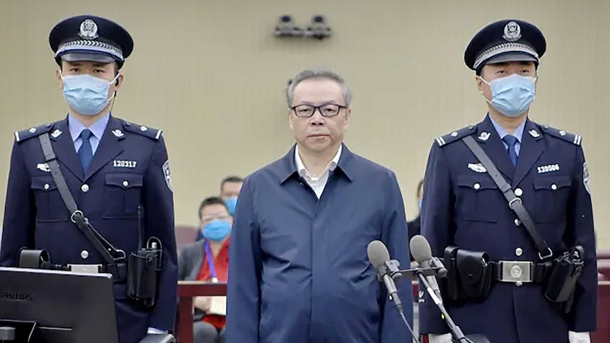 A man in glasses stands between two guards wearing masks.