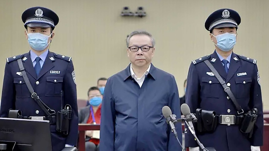A man in glasses stands between two guards wearing masks. A man in glasses stands between two guards wearing masks.