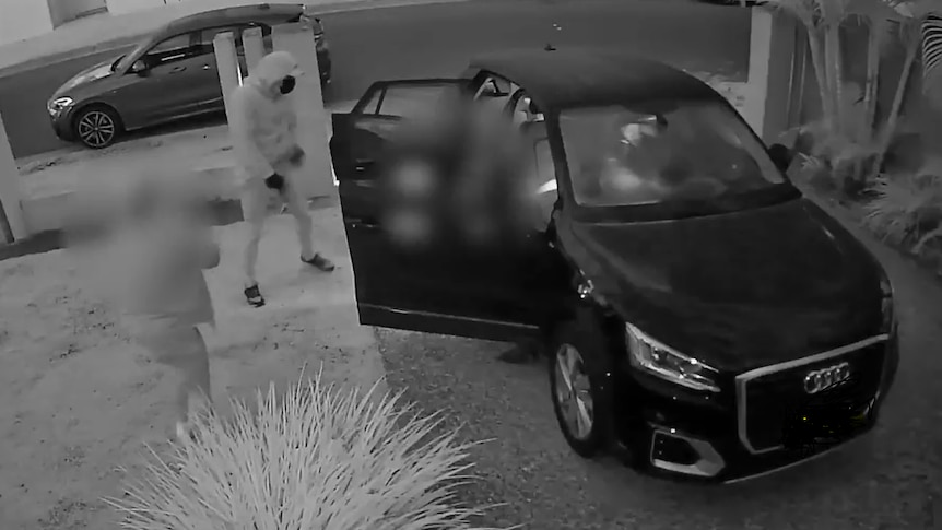 A man in a hooded jumper stands next to a car captured on CCTV