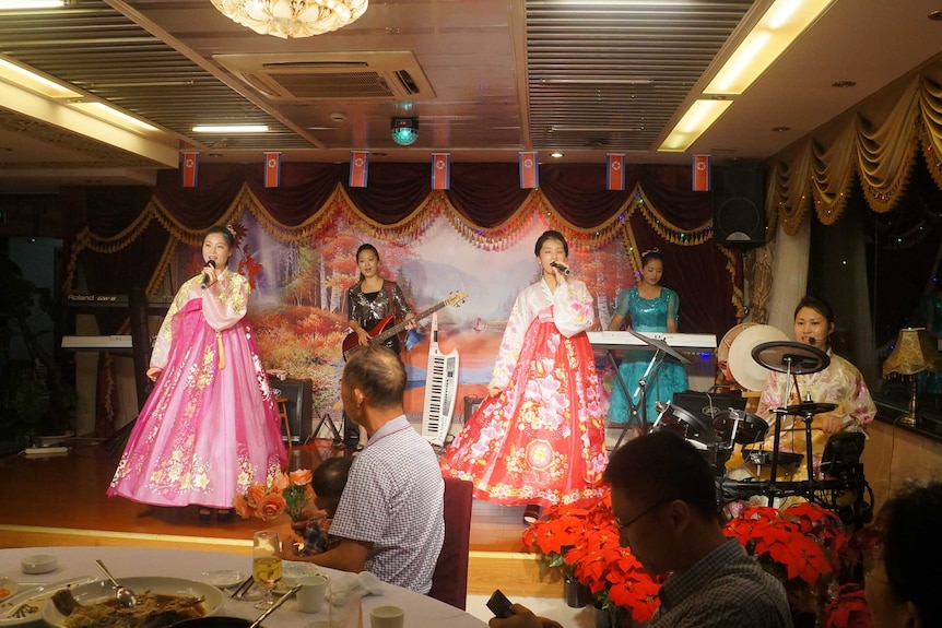 North Korean waitresses in bright traditional clothes perform on a stage.