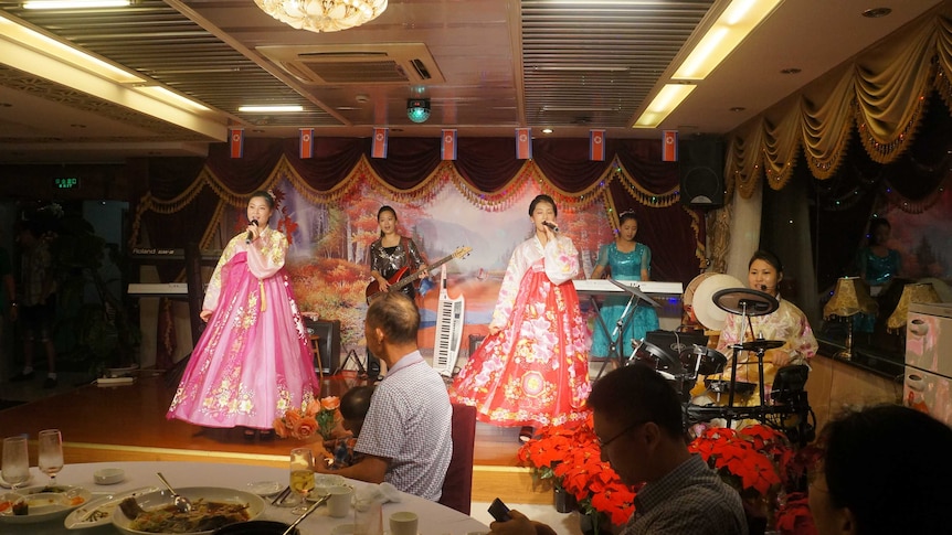 North Korean waitresses in bright traditional clothes perform on a stage.