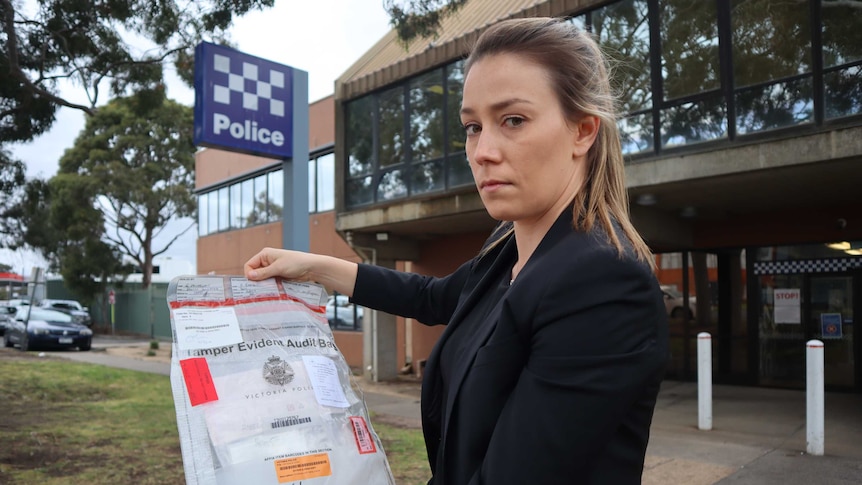 A female police officer out of uniform holds an evidence bag in front of a police station