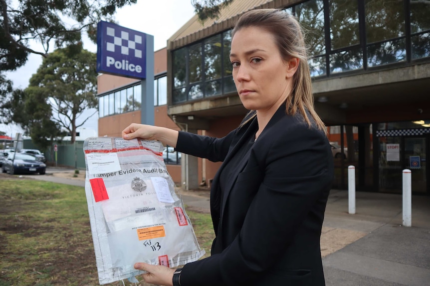 A female police officer out of uniform holds an evidence bag in front of a police station