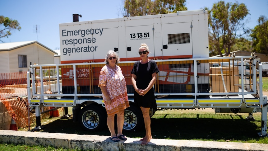 Two women wearing sunglasses stand on a small limestone wall in a grass area, in front of a large generator. The sky is blue.