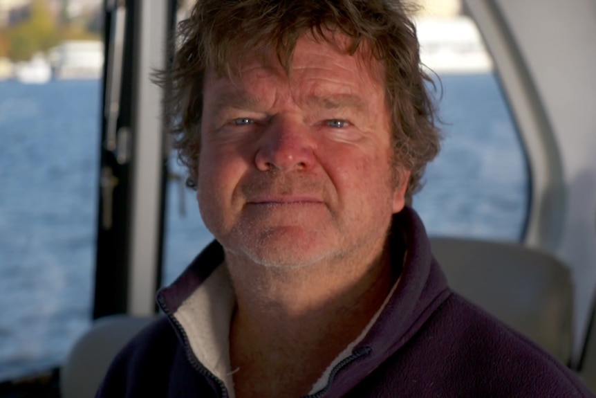 A headshot of a man showing a port window of a small boat and water in the background.