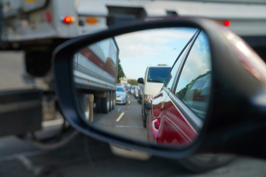 A side car mirror reflects Sydney traffic in peak hour. There are trucks, vans, and cars lined up.