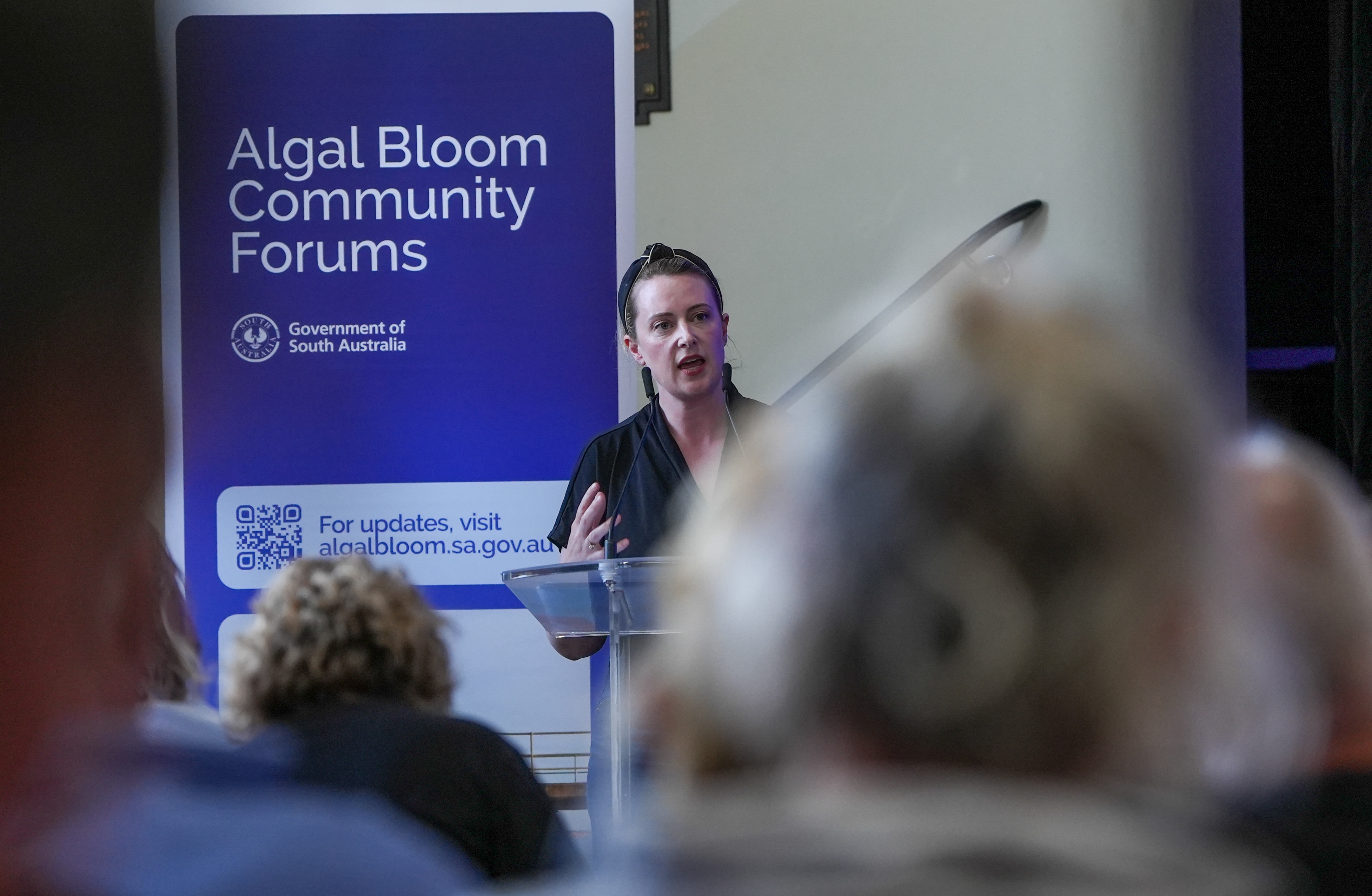 Una mujer habla en un podio junto a un cartel del foro comunitario mientras la gente escucha