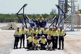 A group of workers in high vis pose smiling in front of machinery from a rocket