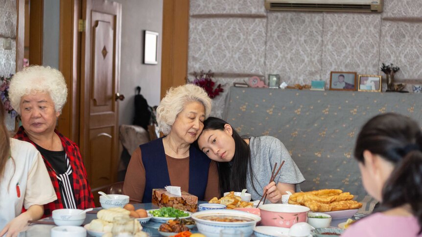 A young Chinese woman rests her head on her grandmother, they sit at a table filled with food