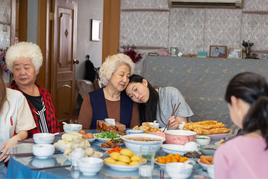 A young Chinese woman rests her head on her grandmother, they sit at a table filled with food