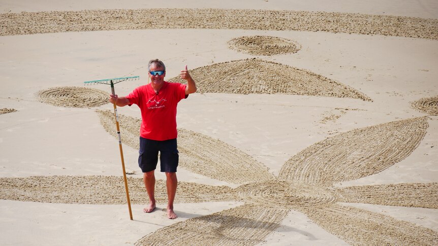 A middle aged man wearing a red t-shirt standing on a beach on a sunny afternoon.